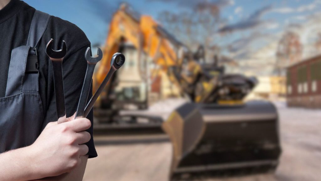 Person i arbetskläder håller tre stora skiftnycklar i fokus, med en grävmaskin och byggarbetsplats i bakgrunden.