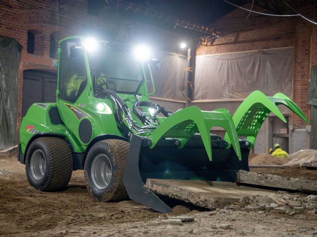 Avant elektrisk kompaktlastare med grip flyttar plank i en inomhusmiljö med tänd arbetsbelysning.
