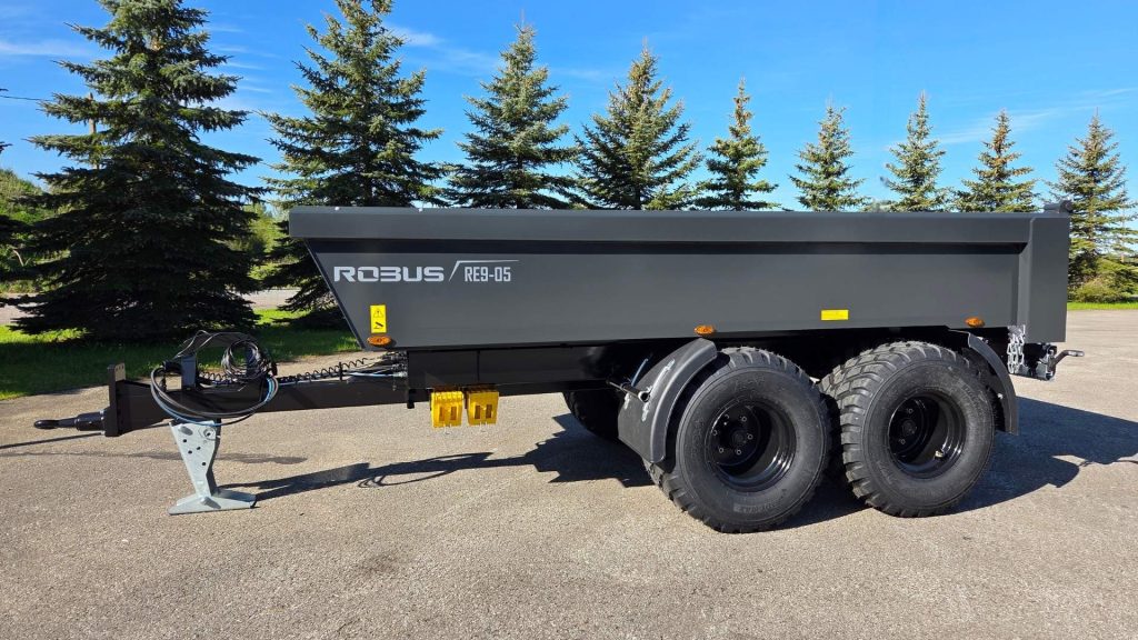 Robus dumpervagn DUMPER RE9-05 i sidovy, svart lack med två kraftiga hjulaxlar, parkerad på asfalt med gröna granar och blå himmel i bakgrunden.