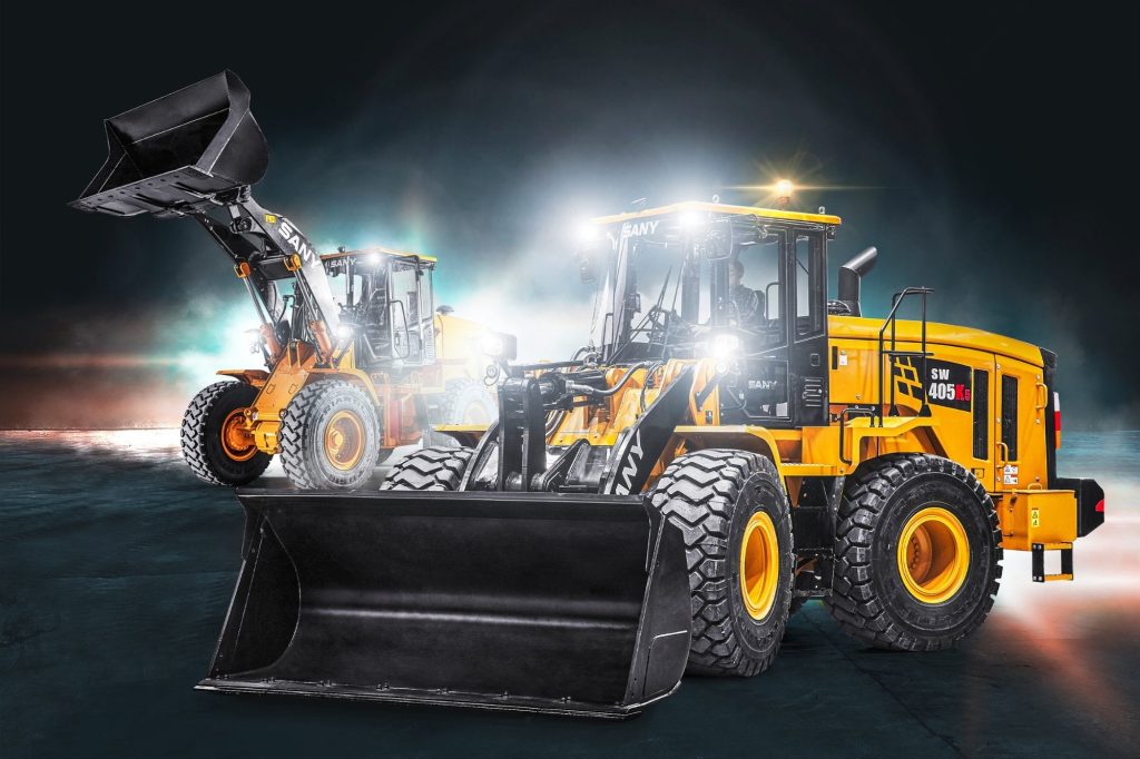 Två stora SANY hjullastare, modell SW405K5, står uppställda inomhus med skoporna i fokus. Den främre maskinen visas rakt framifrån med skopan i markläge, medan den bakre maskinen har skopan upplyft. Bilden har dramatisk belysning och tydligt SANY-varumärke på maskinerna.