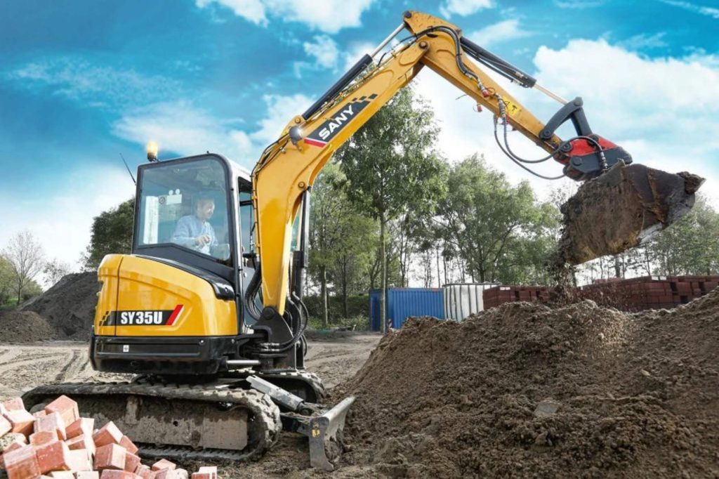 SANY SY35U minigrävare i arbete med grävskopa, lyfter jord på arbetsplats med träd och blå himmel i bakgrunden.